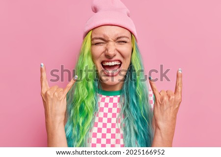Joyful millennial woman makes rock n roll gesture shows she is cool feels very happy has long dyed hair wears hat and dress isolated over pink background. Hipster girl shows heavy metal horn sign