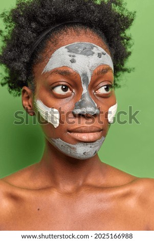 Vertical image of thoughtful curly woman with dark skin looks away has big eyes full lips applies nourishing clay mask on face to remove pores and fine lines poses shirtless against green background