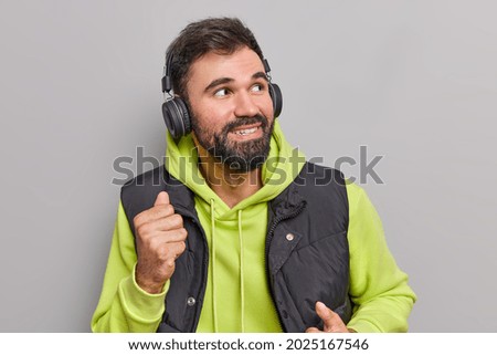 Handsome positive bearded adult man dances carefree moves with rhythm of favorite music looks away wears wireless headphones on ears wears casual hoodie and vest isolated over grey background