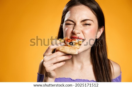 Close up of young woman eating with satisfation, biting pizza slice and looking delighted at camera, try tasty food at pizzeria. Concept of takeaway, fasf-food order or restaurant advertising Royalty-Free Stock Photo #2025171512