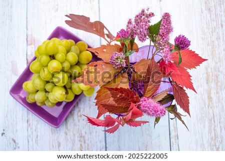 Beautiful bright summer still life. Red grape leaves, clover and pink flowers in a purple vase. In the background, a bunch of green grapes on a light wooden table. Top view. Colorful nature.