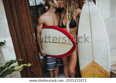 Slender boy and girl kiss. Couple holds surfboards and poses near entrance to house