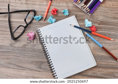 View from above of a workplace concept.  Empty notebook, color pencils, crumpled papers and eyeglasses on the wooden background.  Noise is visible due to the texture of the subjects