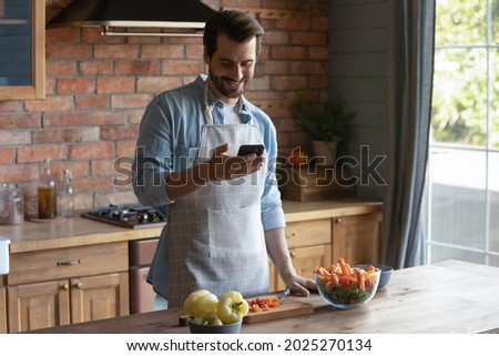 Smiling millennial Caucasian man in apron cook tasty salad for lunch check recipe on smartphone online. Happy young male prepare healthy tasty food for dinner, use cellphone texting on web.