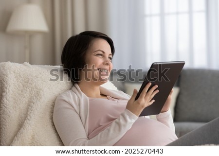 Happy young expecting mom enjoying leisure at home with gadget, reading ebook about childbirth, watching movie on tablet computer, buying baby clothes online, making video call, talking, laughing