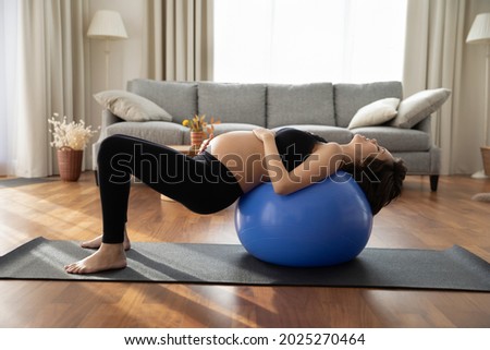 Concentrated pregnant woman doing exercises with fitness ball at home, practicing yoga, pilates with support on fitball, training body, core muscles. Sport activity in pregnancy, prenatal care concept