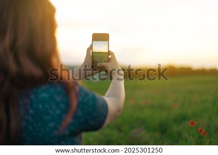 Back view of young girl in a summer dress makes a photo on her smartphone in an autumn field at the golden hour