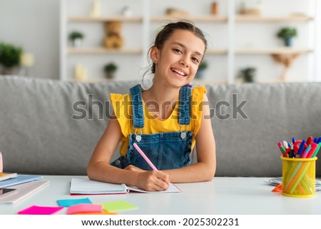 Modern Education Concept. Portrait of smiling little female student sitting at desk in living room, writing notes in her notebook or diary, looking posing at camera, selective focus Royalty-Free Stock Photo #2025302231