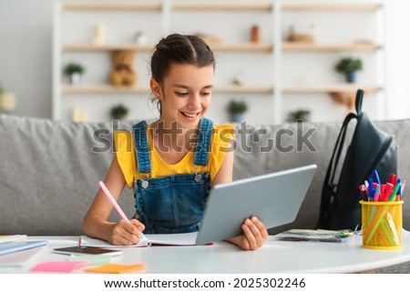 Children and Education Concept. Portrait of positive teen girl using doing homework writing in notebook sitting on couch at desk, holding and using tablet pad computer, enjoying distance homeschooling Royalty-Free Stock Photo #2025302246