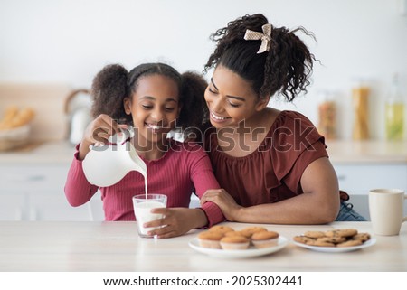 Loving young black mother and teen daughter enjoying fresh homemade cookies, drinking milk, sitting at table in kitchen and smiling, spending time together at home, copy space. Healthy diet concept Royalty-Free Stock Photo #2025302441