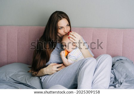 A young long-haired mother sitting in bed in sleepwear hugs and kisses her little daughter.
