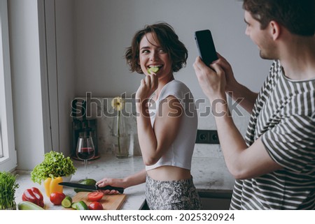 Young couple two woman man 20s in casual t-shirt clothes prepare salad take photo by mobile cell phone eating cucumber slice cook food in light kitchen at home together Healthy diet lifestyle concept