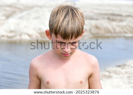 A sad child with his head bowed stands by a small lake on the sand.