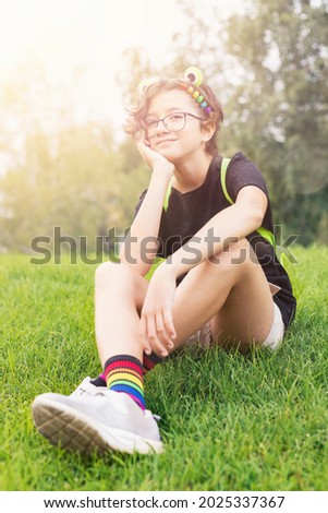 cheerful teenage girl in glasses with colorful hair clips is sitting in meadow on sunny warm day, LGBT-themed, pride