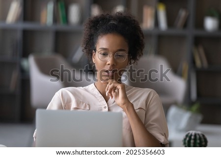 Thoughtful young African American woman in glasses look at laptop screen work online at home office. Pensive ethnic female use computer distant think ponder of problem solution. Technology concept. Royalty-Free Stock Photo #2025356603