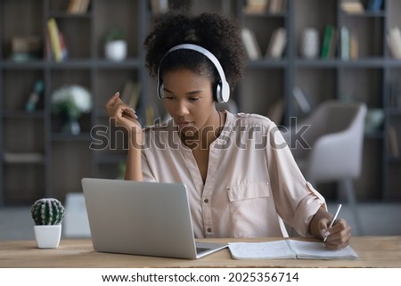 Pensive millennial African American female student in earphones study online on laptop handwrite take notes. Focused young ethnic woman in headphones work distant on computer write summarize in pad. Royalty-Free Stock Photo #2025356714