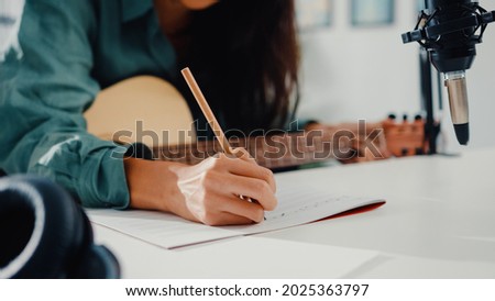 Happy asia woman songwriter play acoustic guitar listen song from smartphone think and write notes lyrics song in paper sit in living room at home studio. Music production at home concept. Royalty-Free Stock Photo #2025363797