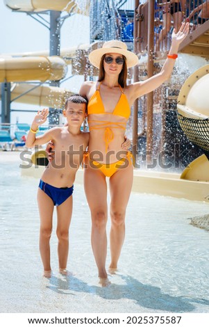 A young mother and son are smiling happily against the background of a larger and modern water park on a sunny day. Happy vacation vacation. Summer holidays and tourism.
