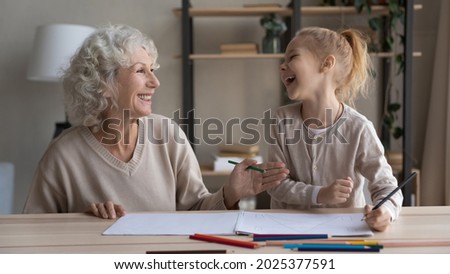 Overjoyed loving old grandmother and small granddaughter have fun drawing painting together in album. Smiling middle-aged Caucasian grandma and little grandchild enjoy hobby art activity at home.