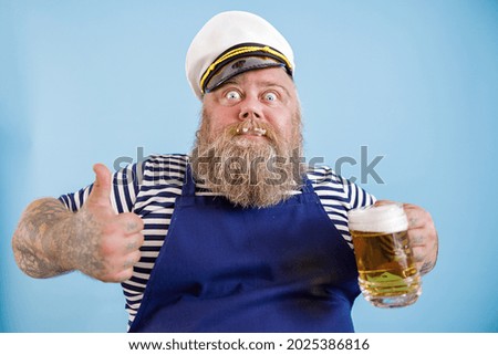 Joyful bearded person with overweight in sailor suit holds mug of tasty beer and shows thumb up on light blue background in studio Royalty-Free Stock Photo #2025386816