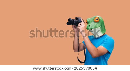 person in frog mask gesture with a camera as a photojournalist looking through the lens on orange background with copy space