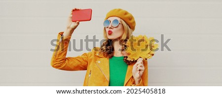 Autumn portrait of beautiful young woman taking selfie by smartphone with yellow maple leaves wearing a french beret on gray background