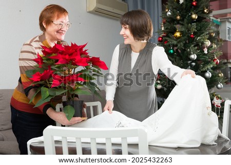 Happy woman with her elderly mother decorating house with white tablecloth and red flowering poinsettia on New Year Eve.. Royalty-Free Stock Photo #2025432263