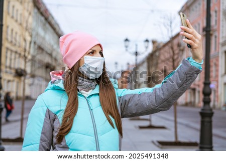 Young woman of Caucasian ethnicity in a warm jacket, in a protective mask, takes pictures on the phone on a cold autumn day