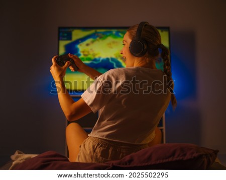 A young girl is playing a video game. She has headphones on her head and a joystick in her hands. She emotionally reacts to what is happening on the screen. Dark background.