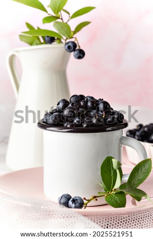 Fresh blueberries in an enamel cup. Pink background. Vintage provance kitchen. Branch of a blueberries, greren leaves. Selective focus (bokeh). Empty space for a text editing. Summer background.