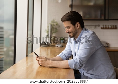 Young man sit at bar counter in office of home kitchen hold smart phone surfing website, chatting on-line, communicate in social media networks, e-date services, modern tech usage, e business concept
