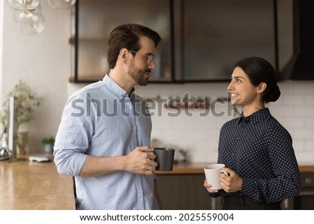 Multiracial colleagues talk during lunch break in office kitchen. Indian woman communicates with workmate hold coffee cups enjoy conversation, discuss work or personal. Good relations at work concept Royalty-Free Stock Photo #2025559004