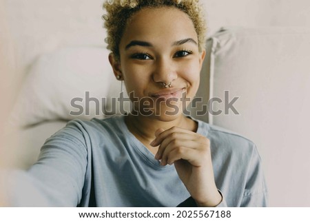 Close-up of cute African ethnicity girl recording stories for her blog using selfie camera of her smartphone, sharing information, dressed in grey blue shirt posing against cosy home background