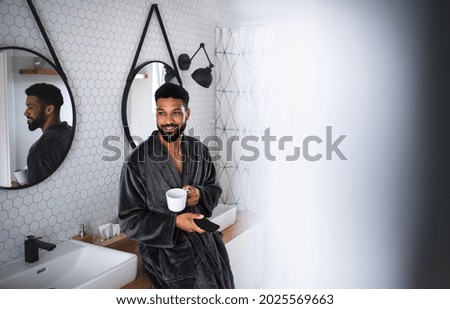 Young man with coffee and bathrobe indoors in bathroom at home, using smartphone. Royalty-Free Stock Photo #2025569663