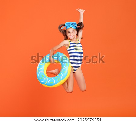 Little funny happy girl in swimsuit and goggles on her head jumping up in air with inflatable ring around waist, isolated over orange studio background. Summertime, vacation an school holidays concept