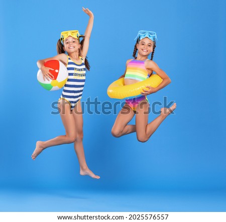 Two funny little girls sisters in swimsuits and swimming goggles jumping up in air with inflatable swimming ring and ball in hands on blue studio background, excited and ready for summer sea vacation Royalty-Free Stock Photo #2025576557