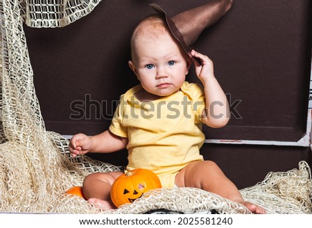 A little girl, dressed as a witch with a hat, sits in a chest and holds a pumpkin. Kid is having fun on Halloween