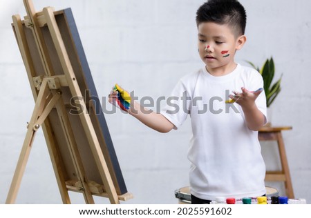 Cute happy little Asian boy painting with watercolor on canvas standing in the white room at home, creative young artist at work. Kids paint. Children draw.