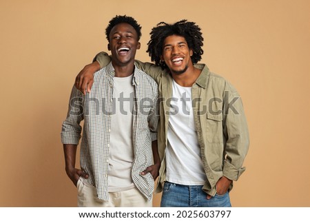 Portrait Of Two Happy Black Guys Embracing While Posing Over Beige Background, Cheerful Young African American Friends In Stylish Clothes Having Fun In Studio, Laughing At Camera, Copy Space Royalty-Free Stock Photo #2025603797