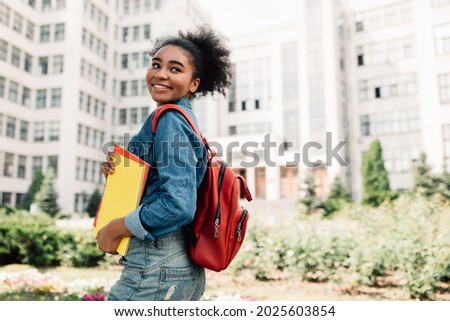 Happy African American Student Girl Posing With Backpack Holding Books Smiling Looking Aside Standing Near University Building Outdoor. Modern Education And Studentship Lifestyle