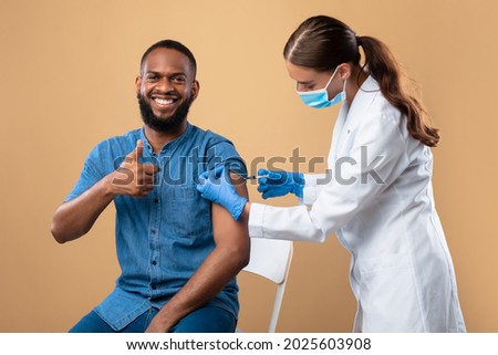 Immunization and health care. Cheerful black male patient receiving covid-19 vaccine injection, showing thumb up on beige studio background. Medical worker vaccinating young man from coronavirus