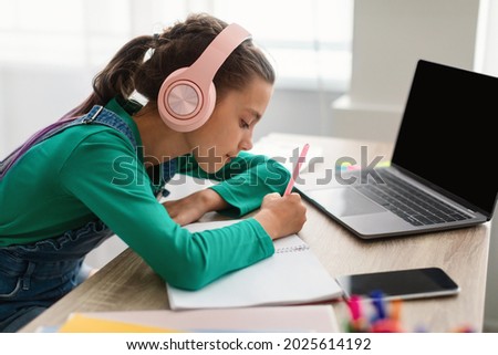 Side view profile of focused little girl writing down information watching webinar on laptop with empty screen or taking notes in her diary, wearing wireless headphones, pc with free space mock up Royalty-Free Stock Photo #2025614192