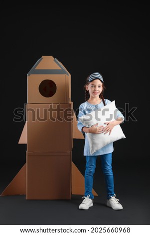 Cute little girl with pillow and cardboard rocket on dark background