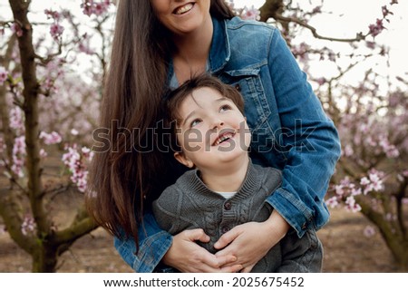 Mother and son picture taken in nature. On a farm with peach pink blossom trees. Hugging and laughter. The mom had beautiful long hair. Mother and son have the same hair. 