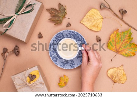 Autumn composition. A female hand holds a cup of coffee, gifts in craft paper, autumn leaves on a beige background. Flat lay, top view