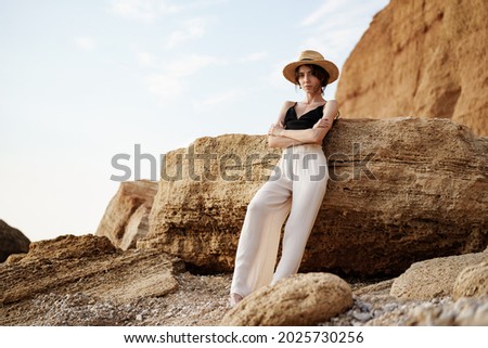 Portrait of woman in black bralette and white trousers leaning on rock at beach Royalty-Free Stock Photo #2025730256