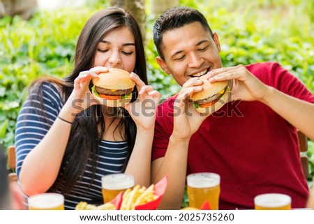 Happy couple eating hamburgers and drinking ice cold beers at an outdoor restaurant in a close up view of them smiling in anticipation as they bite into the food