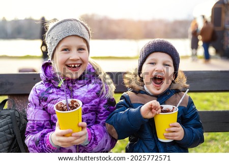 Two cute adorable siblings children sitting on bench drink delicious yummy hot chocolate, tea cocoa from paper cups during walk at city street park or backyard outdoors. Brother and sister enjoy fun