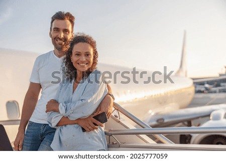 Portrait of happy couple of tourists, man and woman looking excited while standing together outdoors ready for boarding the plane at sunset. Vacation, lifestyle, traveling concept