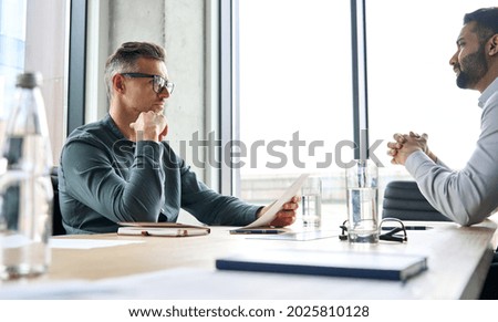 Serious focused mature businessman employer hr or client holding cv or contract listening manager, applicant during job interview, two diverse ethnic partners having negotiations at office meeting. Royalty-Free Stock Photo #2025810128